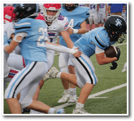 People wearing football uniforms are actively playing on a field with one person holding the ball while another attempts to grab their jersey. Description generated by AI