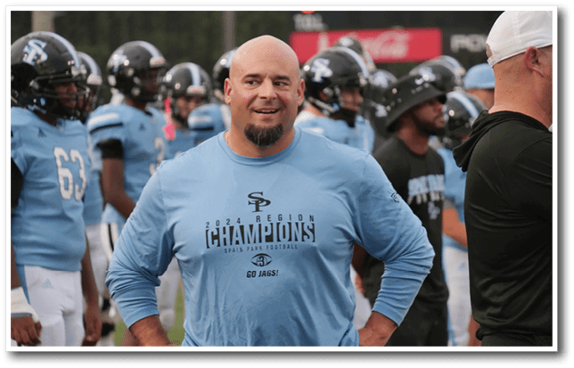 A person wearing a light blue long-sleeve shirt with “2021 Region Champions Spald Park Football Go Jags!" stands smiling with hands on hips on a football field surrounded by players in light blue and white uniforms. Description generated by AI