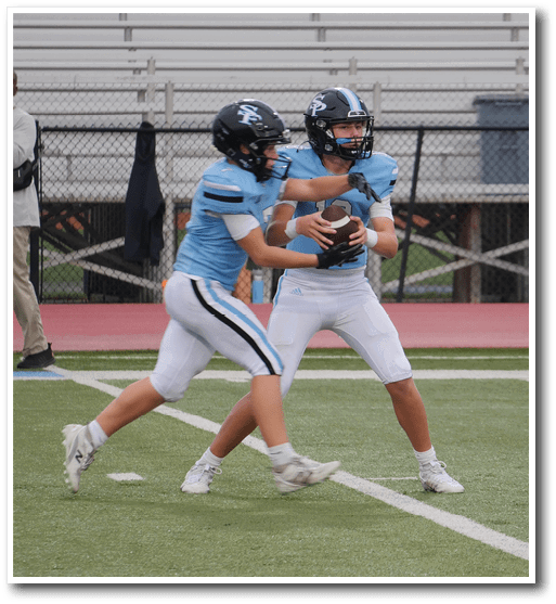 Two people in light blue football uniforms and helmets are on a field with one handing a football to the other. Description generated by AI