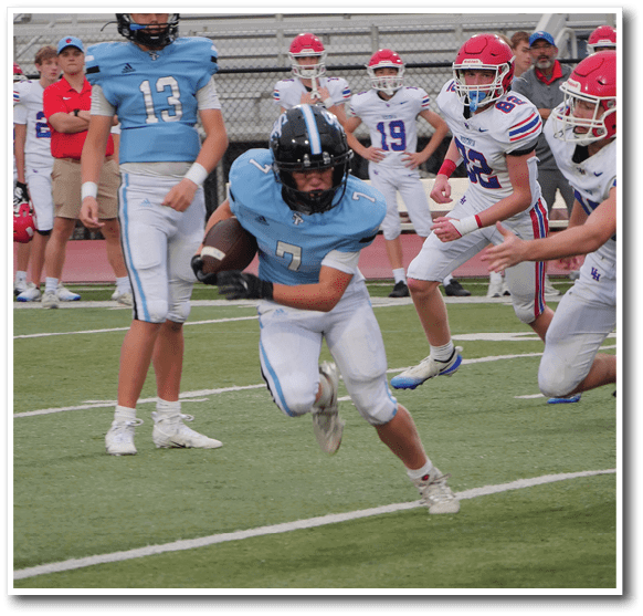A person in a light blue football uniform is running with the ball on a field while other players in white and blue uniforms are nearby. Description generated by AI