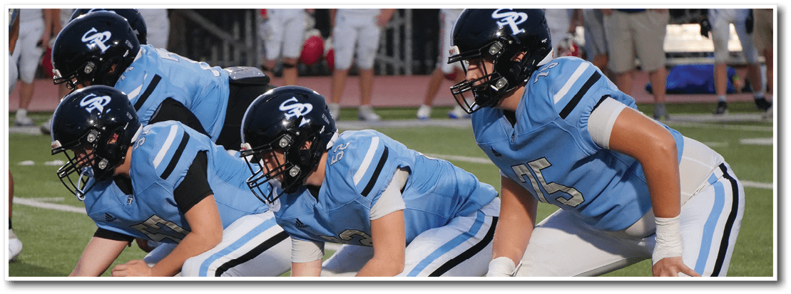 Four people in blue football uniforms and helmets are crouched in a ready position on a grassy field during a game. Description generated by AI