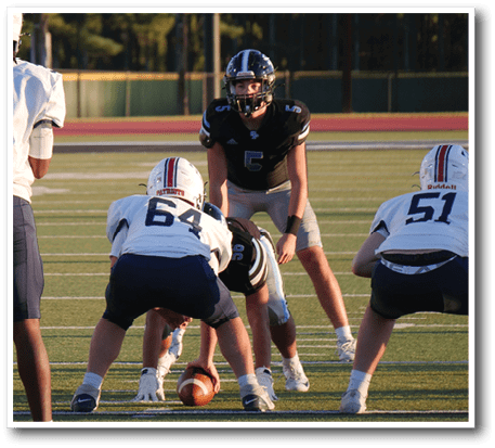 A group of people in football uniforms are positioned on a field with a scoreboard in the background showing one team leading. Description generated by AI