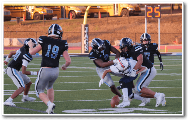 Several people in football uniforms are actively engaged in a tackle on a football field with a referee and a scoreboard visible in the background. Description generated by AI
