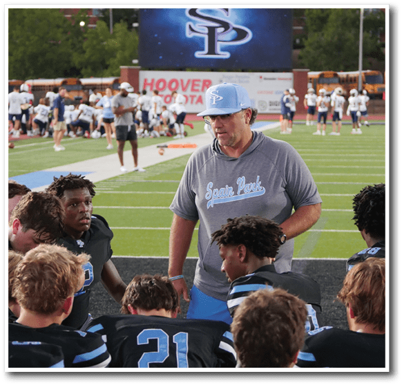 A group of people wearing sports uniforms and a person in a cap and hoodie are on the sidelines of a football field with players and school buses in the background. Description generated by AI