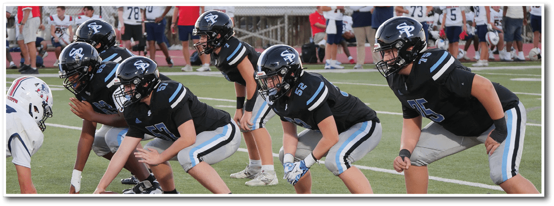 Five people in black football uniforms and helmets are crouched in a ready position on a football field facing opponents in white uniforms. Description generated by AI