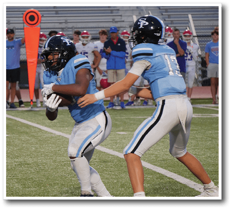 Two people in light blue football uniforms and helmets are on a football field with one handing off the ball to the other. Description generated by AI