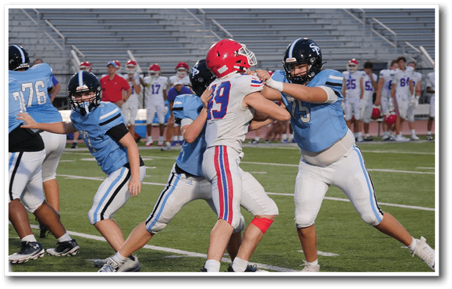 People wearing football uniforms are engaged in a play on a football field with one player in a red helmet being blocked by two players in blue jerseys. Description generated by AI