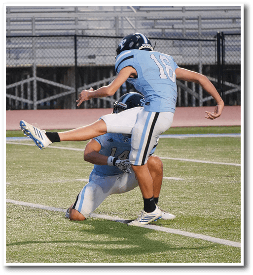 Two people in light blue football uniforms are on a field with one person holding the ball and kneeling while the other is kicking the ball. Description generated by AI