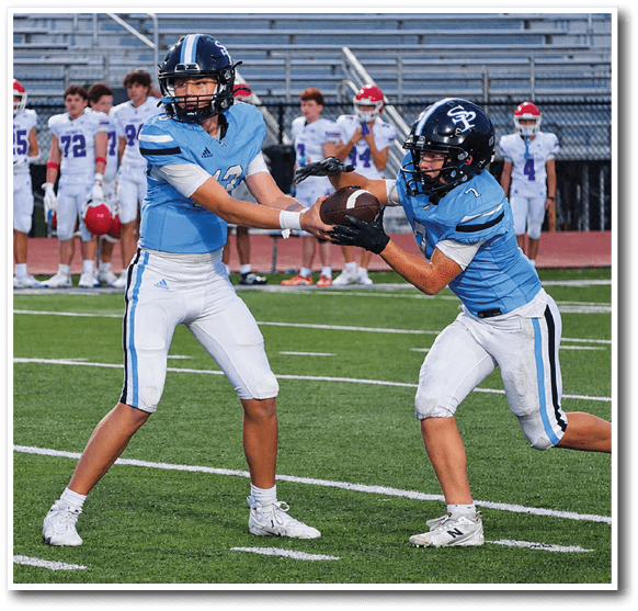 Two people in blue football uniforms are on a field with one handing a football to the other while players in white uniforms stand in the background. Description generated by AI