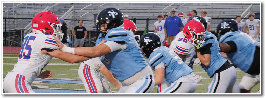 People in football uniforms are engaged in a play on a field with some players blocking and others preparing to move. Description generated by AI