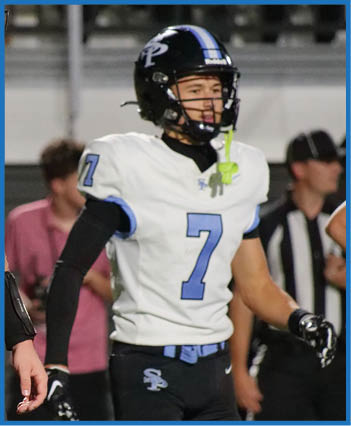 A person wearing a white and blue football uniform with the number seven and a black helmet stands on a field with other people in the background. Description generated by AI