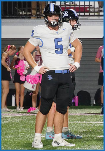 Two people in football uniforms stand on a field with their hands on their hips while spectators watch from the bleachers behind them. Description generated by AI