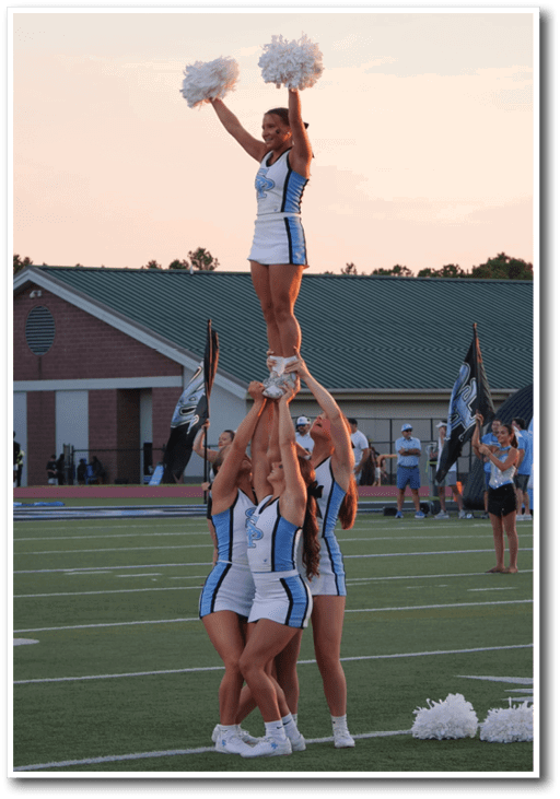 Adults in cheerleading uniforms perform a stunt on a sports field with a building and inflatable tunnel in the background during a sunset. Description generated by AI