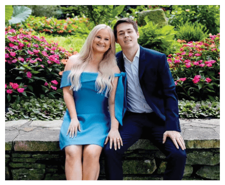 Two adults dressed formally are sitting closely together on a stone bench in a garden with vibrant flowers and greenery. Description generated by AI