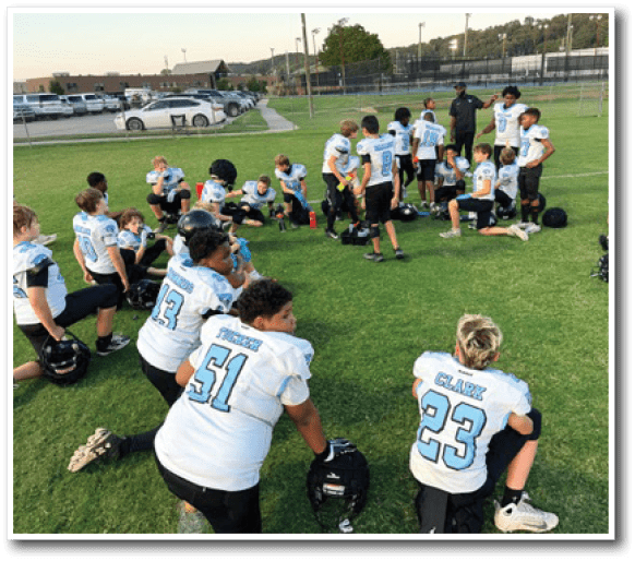 A group of young people in football uniforms are gathered on a grassy field, some sitting and some standing, appearing to listen to a coach during practice or a game break. Description generated by AI