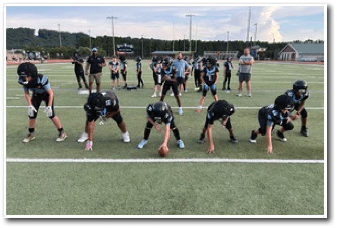 A group of people in football uniforms are lined up on a field in a ready stance with coaches and other team members standing behind them. Description generated by AI