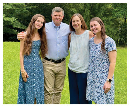 Four adults stand closely together on grass wearing casual clothing including two floral dresses, pants, and a belt. Description generated by AI