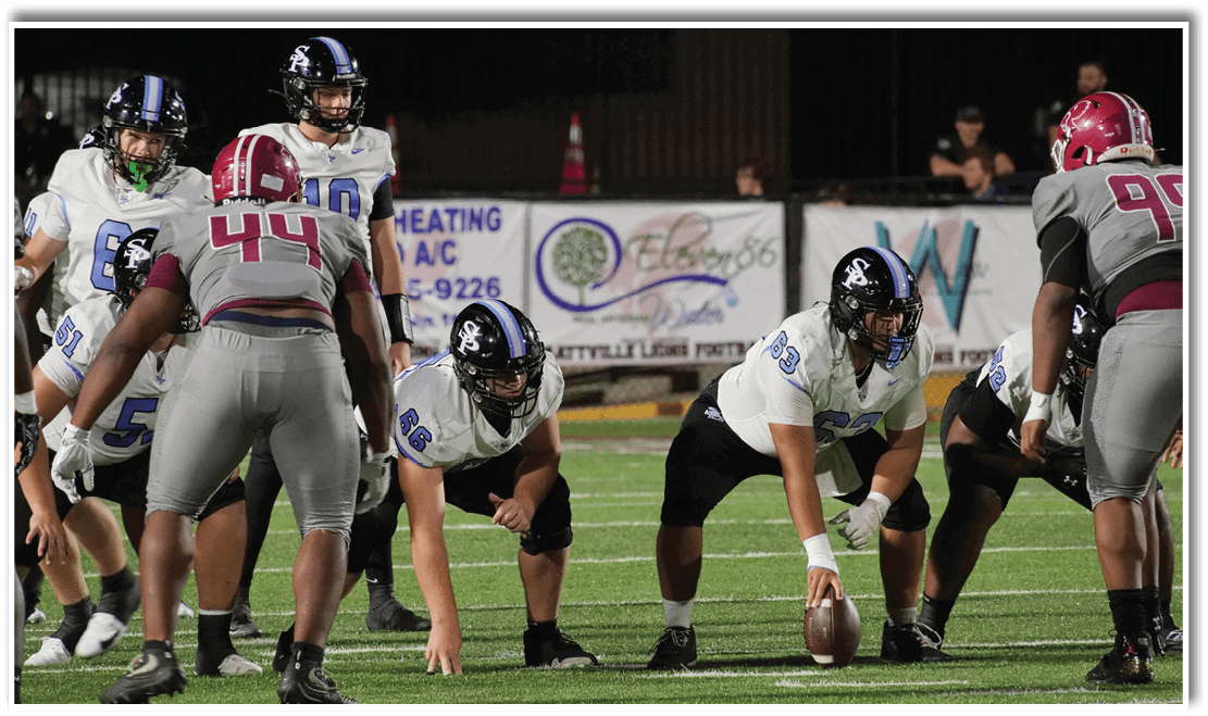 Several people in football uniforms are positioned on a field, preparing for a play during a game. Description generated by AI