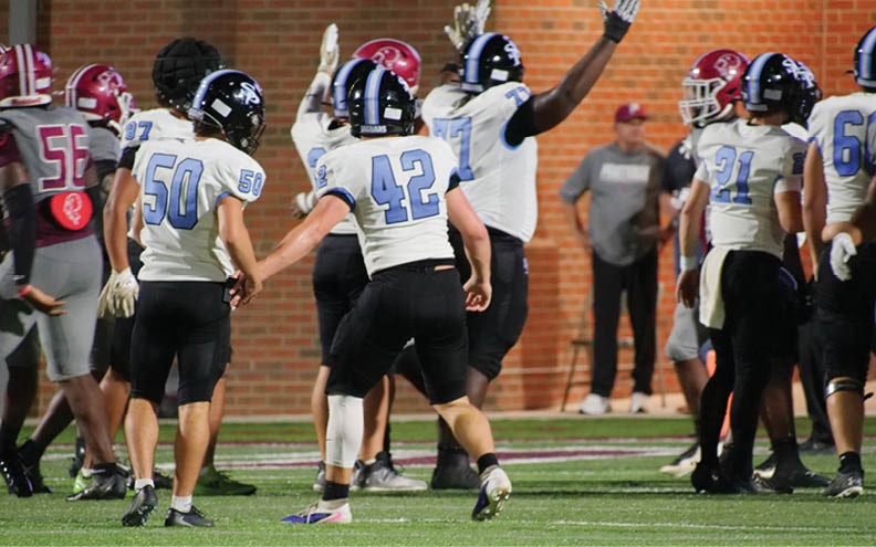 People wearing football uniforms from two different teams are gathered on a field during a game with one person raising an arm. Description generated by AI