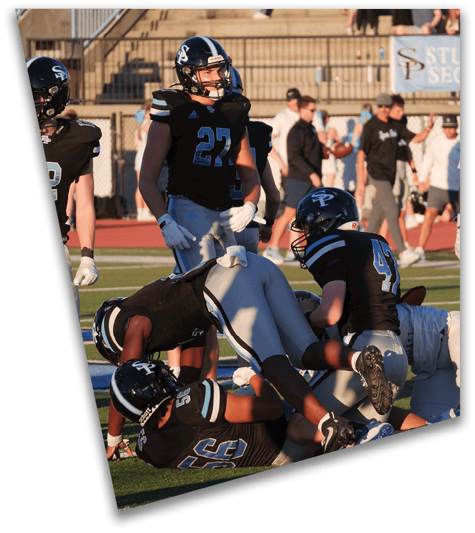 Players in black and white football uniforms are engaged in a tackle on a grassy field with spectators in the background. Description generated by AI