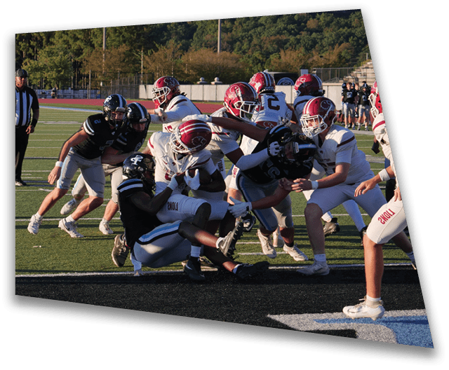 A group of people wearing football helmets and uniforms are engaged in a play on a football field with a referee observing in the background. Description generated by AI