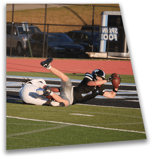 Two people in football uniforms are on the ground with one reaching out to catch a football while a referee watches nearby on a sports field. Description generated by AI