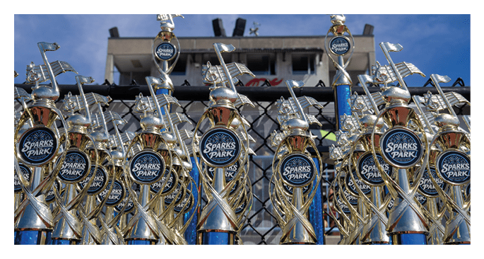 Multiple shiny trophies with musical note designs and “Sparks Park" labels are displayed in front of a building under a clear blue sky. Description generated by AI