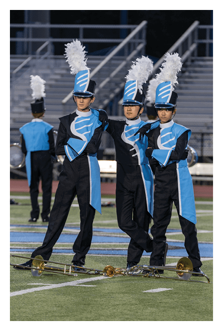 Three people in blue and black marching band uniforms with tall hats adorned with white plumes stand closely together on a field. Description generated by AI
