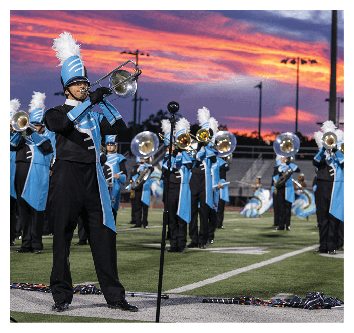 A marching band dressed in blue and black uniforms with white plumes is performing on a football field at sunset. Description generated by AI
