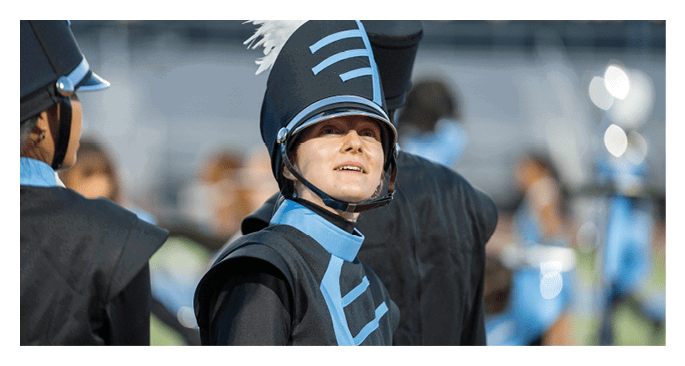 A person wearing a blue and black marching band uniform with a tall hat and white plume is looking slightly upward while standing among other similarly dressed people. Description generated by AI