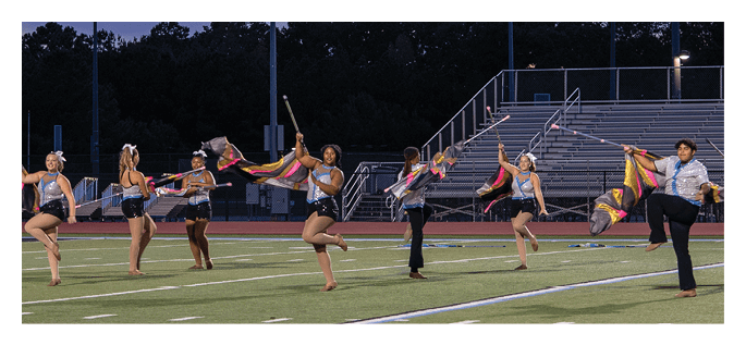 A group of people dressed in colorful uniforms perform a synchronized flag routine on a sports field at dusk. Description generated by AI