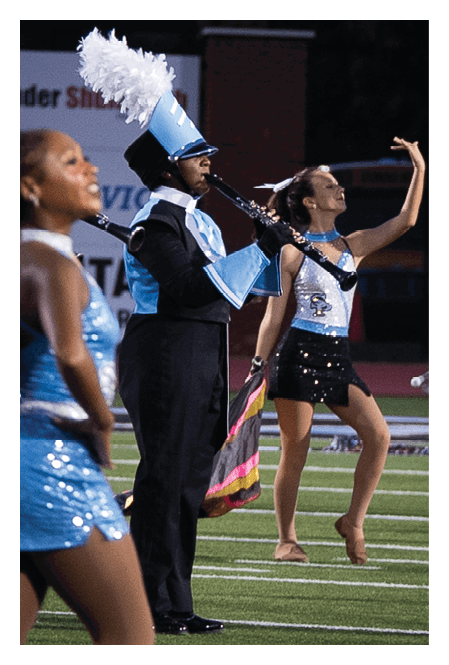 People in a marching band uniform and sequined dance costumes perform on a football field at night. Description generated by AI