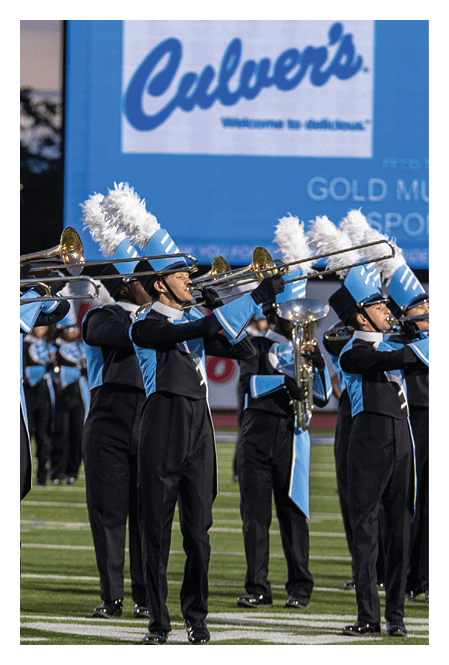 People in blue and black marching band uniforms with white plumed hats are playing trombones on a field with a large Culver's advertisement in the background. Description generated by AI