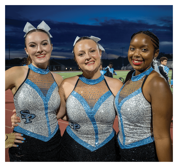 Three adults wearing sparkly blue and silver dance or cheer uniforms with white bows in their hair are smiling and posing together on a sports field at dusk. Description generated by AI
