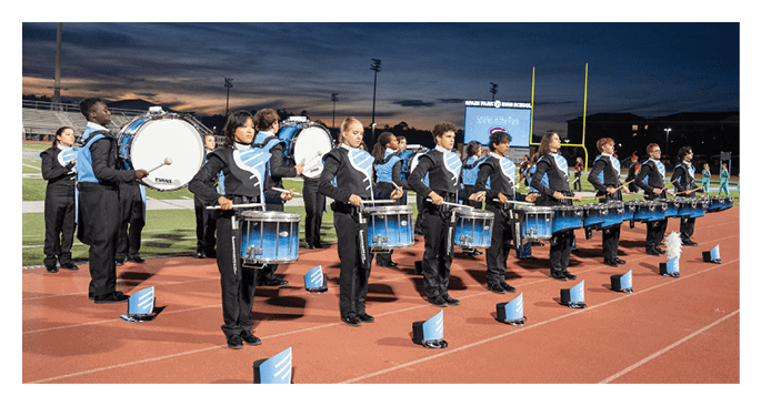 A group of people in matching uniforms are standing in a line on a track field playing drums during an evening outdoor event. Description generated by AI
