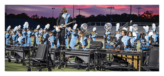 A marching band dressed in blue and black uniforms performs on a field at sunset with one person standing on a platform playing an electric guitar. Description generated by AI