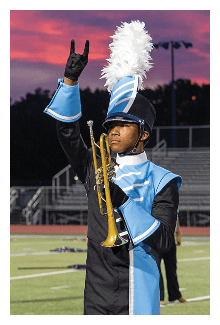 A person in a blue and black marching band uniform with a tall white plume on the hat is holding a trumpet and raising one hand with fingers forming a gesture against a sunset sky background. Description generated by AI