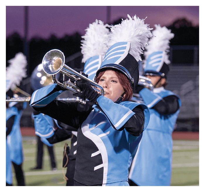 People wearing blue and white marching band uniforms with tall feathered hats are playing brass instruments at sunset. Description generated by AI