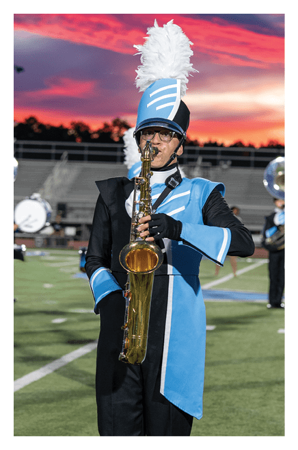 A person in a blue and black marching band uniform with a tall feathered hat is playing a saxophone on a football field at sunset. Description generated by AI