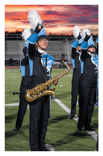 A person in a blue and black marching band uniform with a feathered hat is playing a saxophone on a field at sunset. Description generated by AI