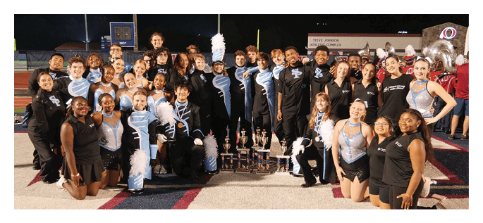 A group of people dressed in marching band uniforms and casual black outfits kneel on a field around several trophies. Description generated by AI