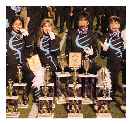 Four people in matching band uniforms are kneeling on a field, holding medals in their mouths and surrounded by numerous trophies and plaques. Description generated by AI