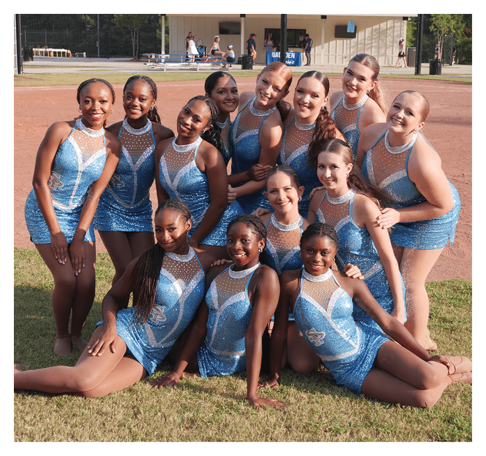 A group of people wearing matching sparkly blue and white dance costumes pose closely together outdoors on grass with a sports field in the background. Description generated by AI