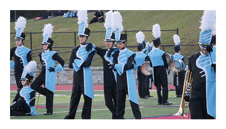People in blue and black marching band uniforms with tall white plumes stand and sit on a field with musical instruments scattered around. Description generated by AI