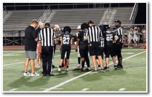 People in sports uniforms and referees are gathered on a football field near empty bleachers during dusk. Description generated by AI