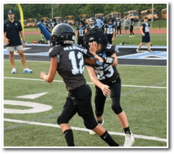 Children in football uniforms are playing on a field with a coach and other players in the background near a goalpost. Description generated by AI
