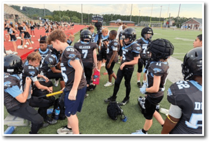 Children wearing football uniforms and helmets are gathered on the sidelines of a sports field with a running track and cheerleaders in the background. Description generated by AI
