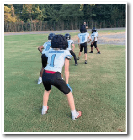 Children wearing football helmets and uniforms are lined up on a grassy field, preparing for a play or drill during practice. Description generated by AI