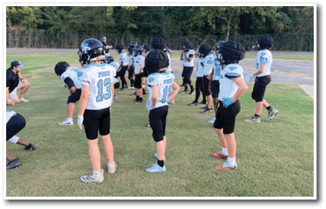 A group of children wearing football helmets and uniforms stand on a grassy field, appearing to prepare for practice or a game. Description generated by AI