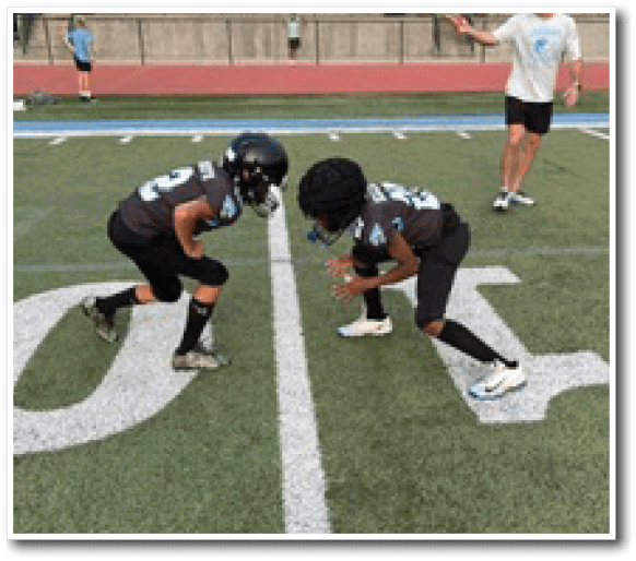 Two children wearing football helmets and uniforms are crouched facing each other on a marked sports field with an adult standing nearby. Description generated by AI